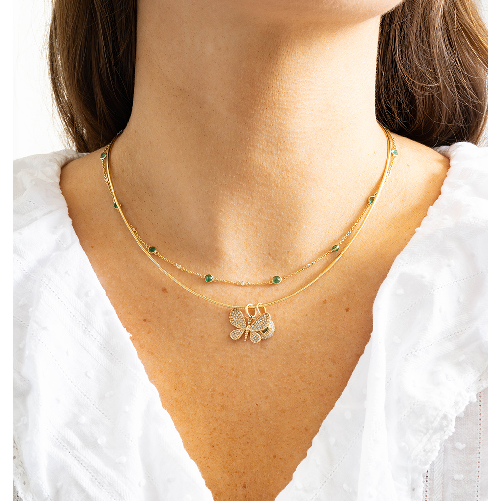 A close-up of a model's neck and chest wearing a delicate double-layered gold necklace. The top layer features small round green gemstones spaced along the chain, while the bottom layer has a textured gold pendant shaped like a butterfly with tiny white stones inlaid inside its wings.