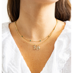A close-up of a model's neck and chest wearing a delicate double-layered gold necklace. The top layer features small round green gemstones spaced along the chain, while the bottom layer has a textured gold pendant shaped like a butterfly with tiny white stones inlaid inside its wings.
