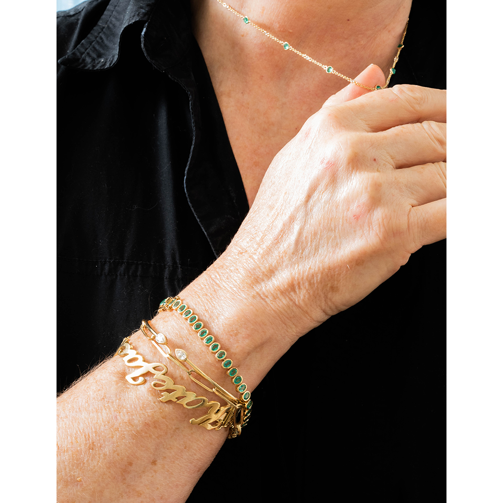 A close-up of a person's wrist wearing multiple gold bracelets, including a nameplate bracelet, a green stone bracelet, and a slim gold chain, with a matching delicate necklace also visible.