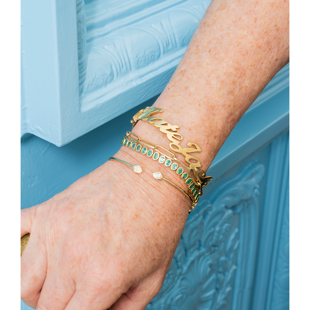 A close-up of a person's hand and wrist wearing multiple gold bracelets, including a script name bracelet and a bracelet with green stones, against a light blue paneled background.