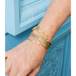 A close-up of a person's hand and wrist wearing multiple gold bracelets, including a script name bracelet and a bracelet with green stones, against a light blue paneled background.