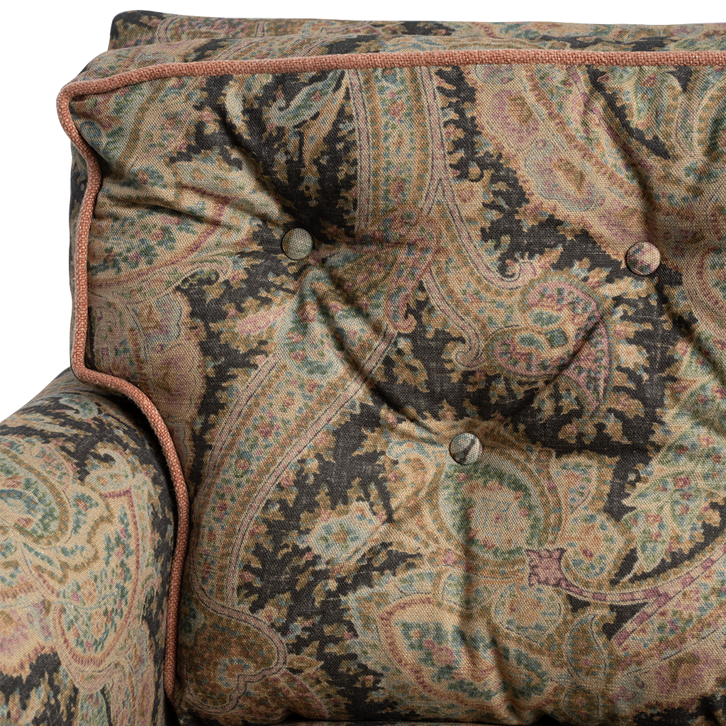 Close-up of a tufted armchair backrest and arm upholstered in an ornate paisley fabric with a rust-colored braided piping trim.
