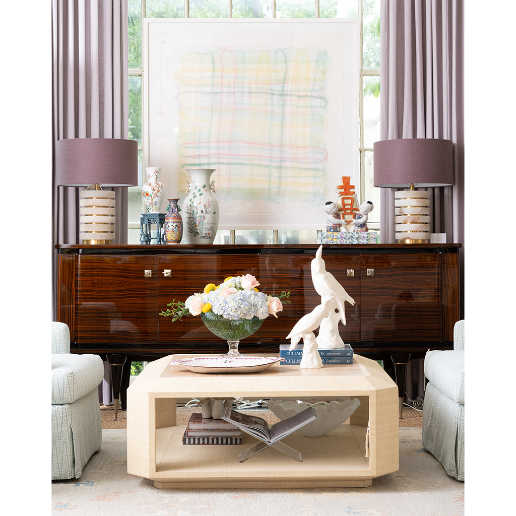 A light natural wood rectangular coffee table with open lower shelves, styled in a bright living room in front of a dark wood sideboard and between two upholstered armchairs.