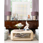 A light natural wood rectangular coffee table with open lower shelves, styled in a bright living room in front of a dark wood sideboard and between two upholstered armchairs.