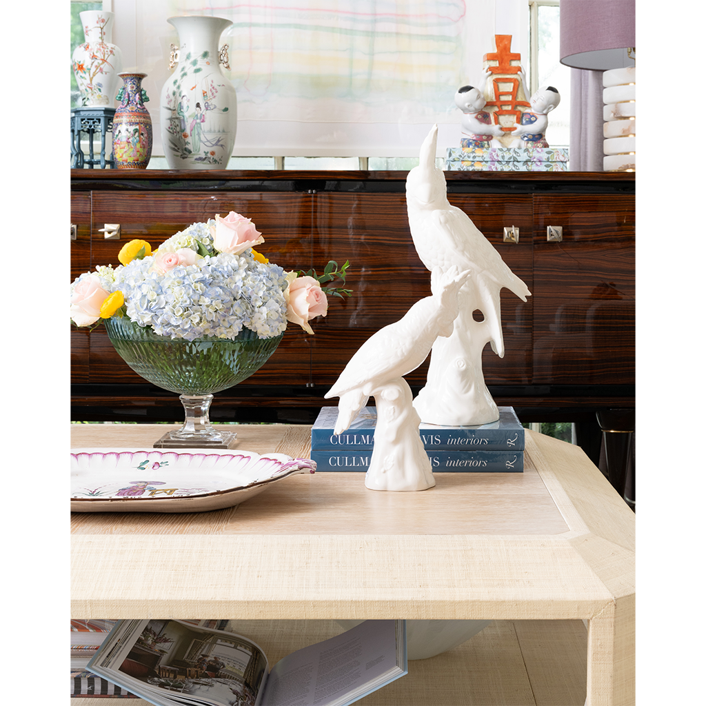 A pair of white ceramic bird sculptures displayed on stacked blue books on a light wood table, with a glass vase of flowers and decorative vases in the background.