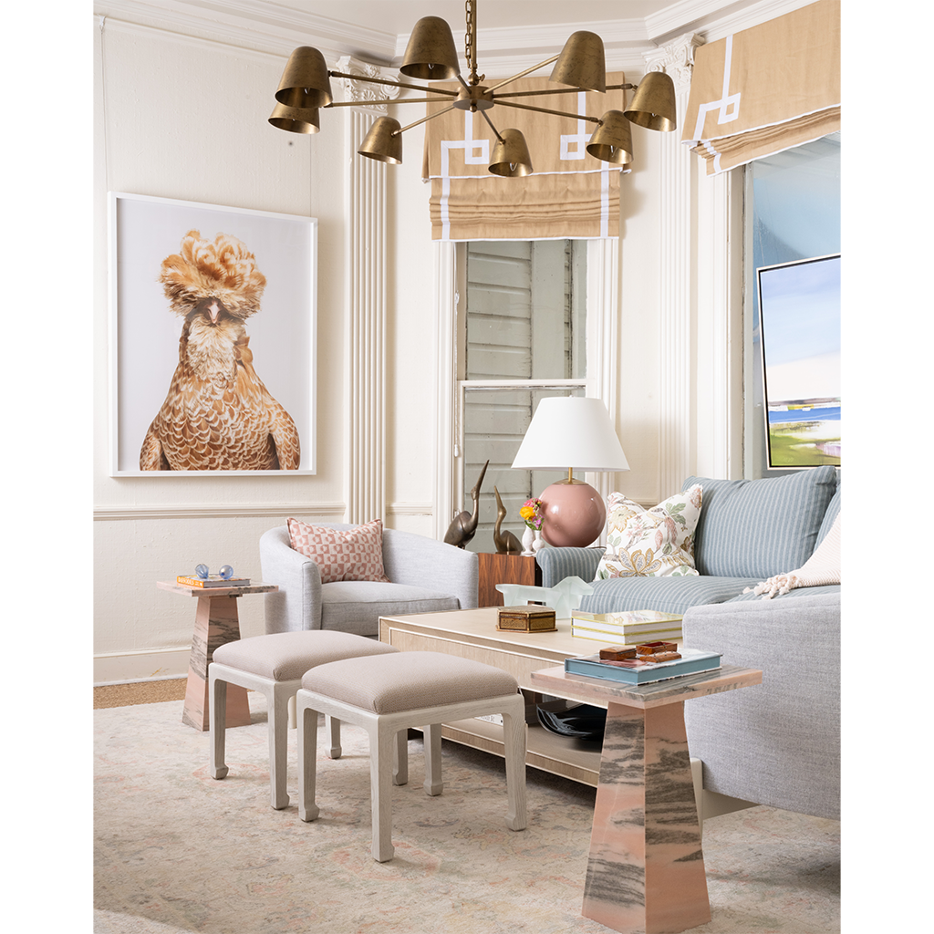 A styled living room interior featuring a large framed art print of a rooster on the wall above a light gray armchair. The room includes a light blue sofa, a wooden coffee table, a chandelier, and a window with beige blinds.