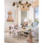 A styled living room interior featuring a large framed art print of a rooster on the wall above a light gray armchair. The room includes a light blue sofa, a wooden coffee table, a chandelier, and a window with beige blinds.