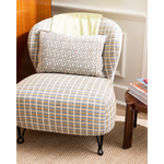 A classic wingback armchair upholstered in a beige, blue, and white plaid fabric, styled with a matching patterned lumbar pillow and a pale yellow throw blanket. The chair is set in a cozy room next to a small wooden side table.
