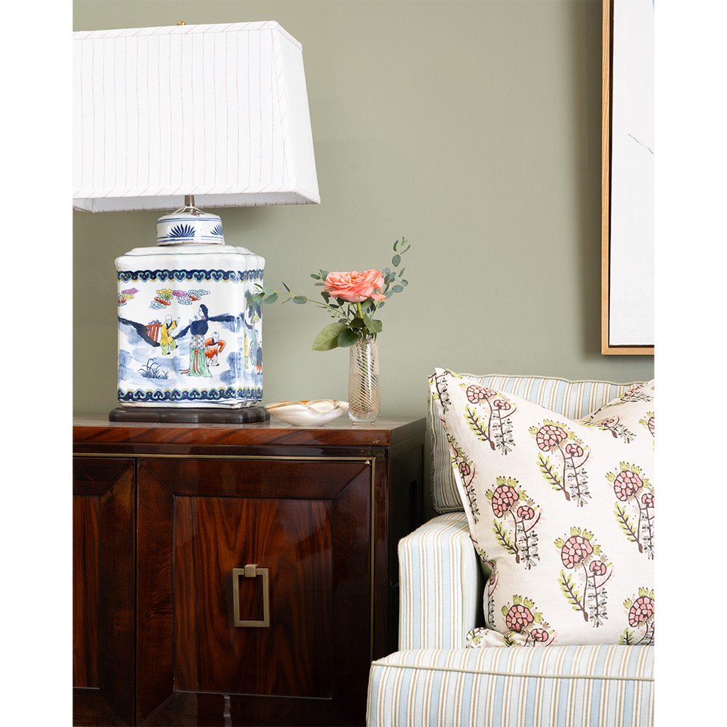 Decorative lamp on a wooden side table with a vase of flowers and a floral-patterned pillow on a sofa.