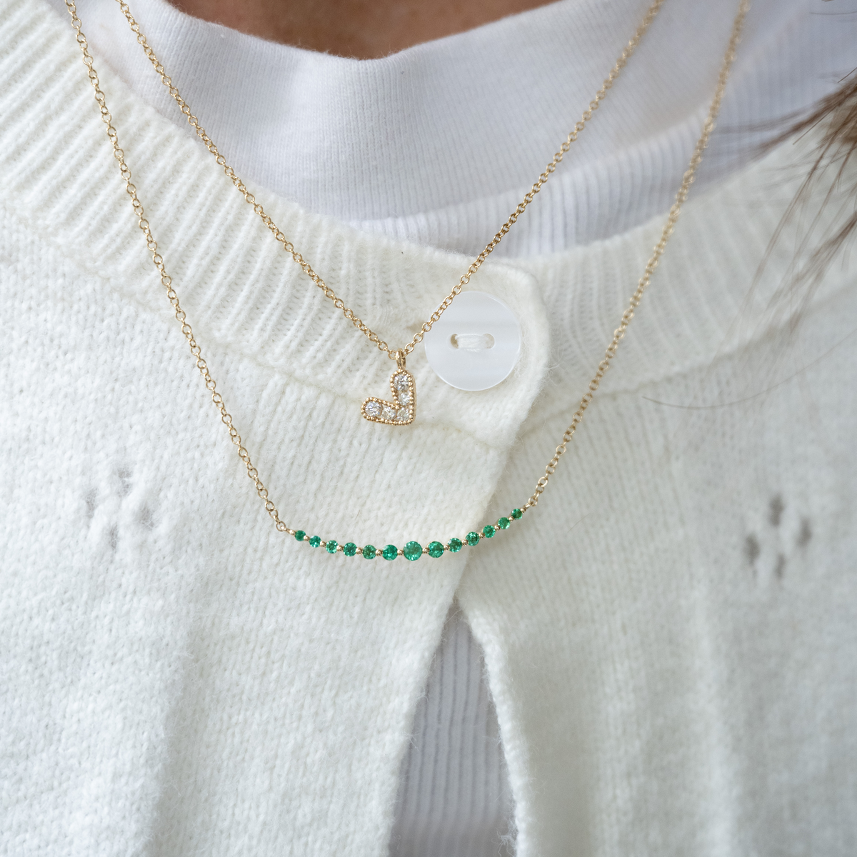 Two gold necklaces with a heart pendant and green gemstones on a white sweater.