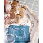 A styled tabletop scene with a decorative rectangular tray holding four metallic gold shot glasses, a folded blue napkin with white embroidered border, and part of a clear drinking glass. The tray has a leaf pattern and sits on a blue plaid tablecloth.