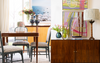 A game table and chairs next to a wooden sideboard with art, books, a lamp, and two pieces of art.