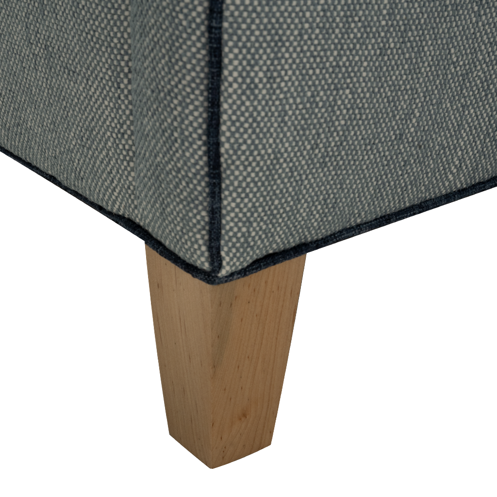 Close-up of the corner of a upholstered furniture piece with a textured blue fabric and dark piping, showing a light natural wood leg on a white background.