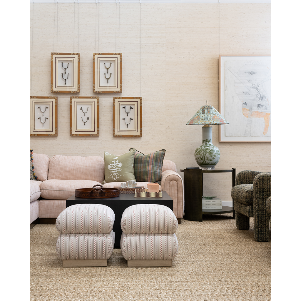 A styled living room scene with a blush pink sofa, two matching upholstered ottomans, a dark wood coffee table, framed wall art, a patterned ceramic lamp, and a textured armchair against a light beige wall.