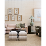 A styled living room scene with a blush pink sofa, two matching upholstered ottomans, a dark wood coffee table, framed wall art, a patterned ceramic lamp, and a textured armchair against a light beige wall.