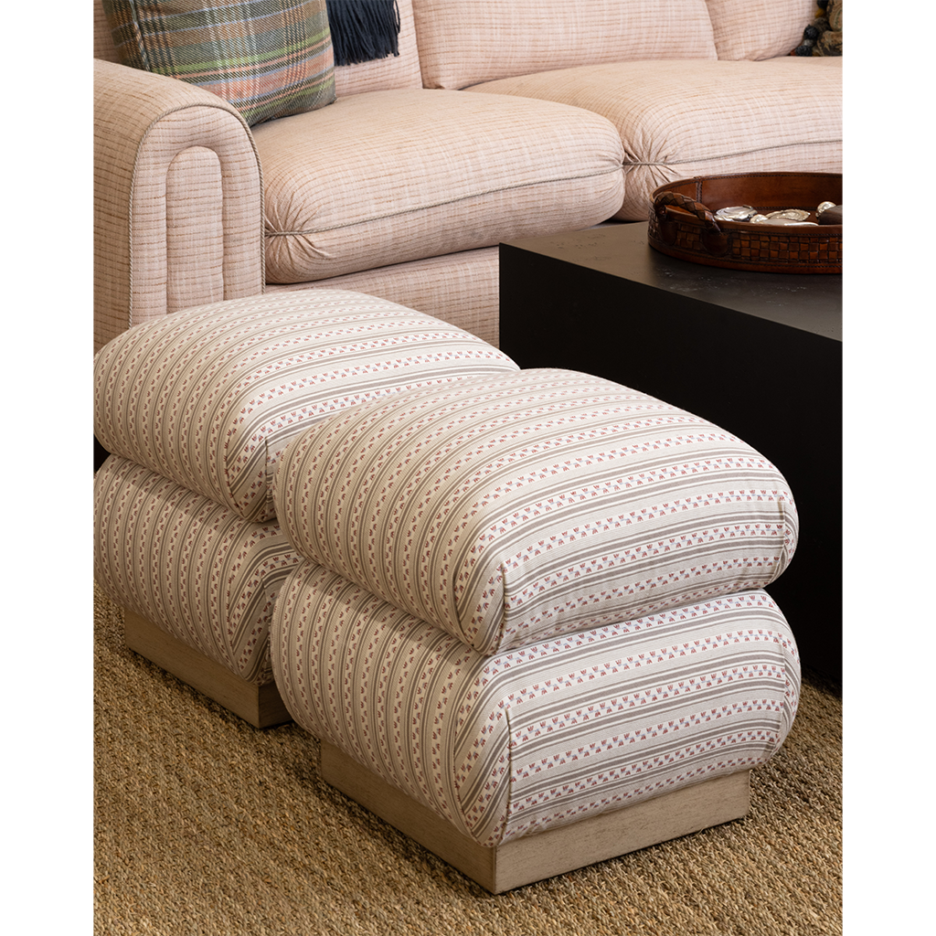 A two-seat upholstered ottoman with plush cushions and a striped pattern in beige, cream, and brown, shown in a living room setting next to a matching sofa and a black coffee table.