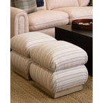 A two-seat upholstered ottoman with plush cushions and a striped pattern in beige, cream, and brown, shown in a living room setting next to a matching sofa and a black coffee table.
