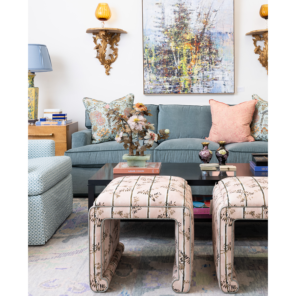 A styled living room scene featuring two upholstered ottomans with a beige floral pattern in the foreground, a blue-gray sofa with patterned and pink pillows in the background, and decorative wall sconces and framed art.