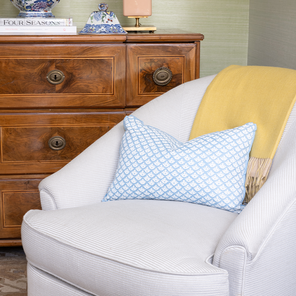 Gray armchair with a blue patterned pillow and yellow blanket in front of a wooden dresser.