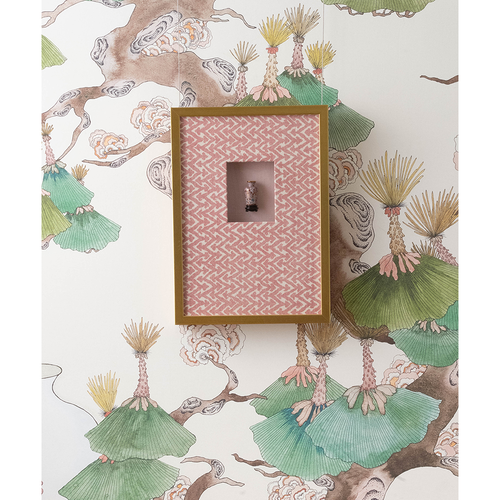 A framed decorative wall piece with a pink patterned insert and a small central figure, mounted against wallpaper featuring green and beige botanical branches.