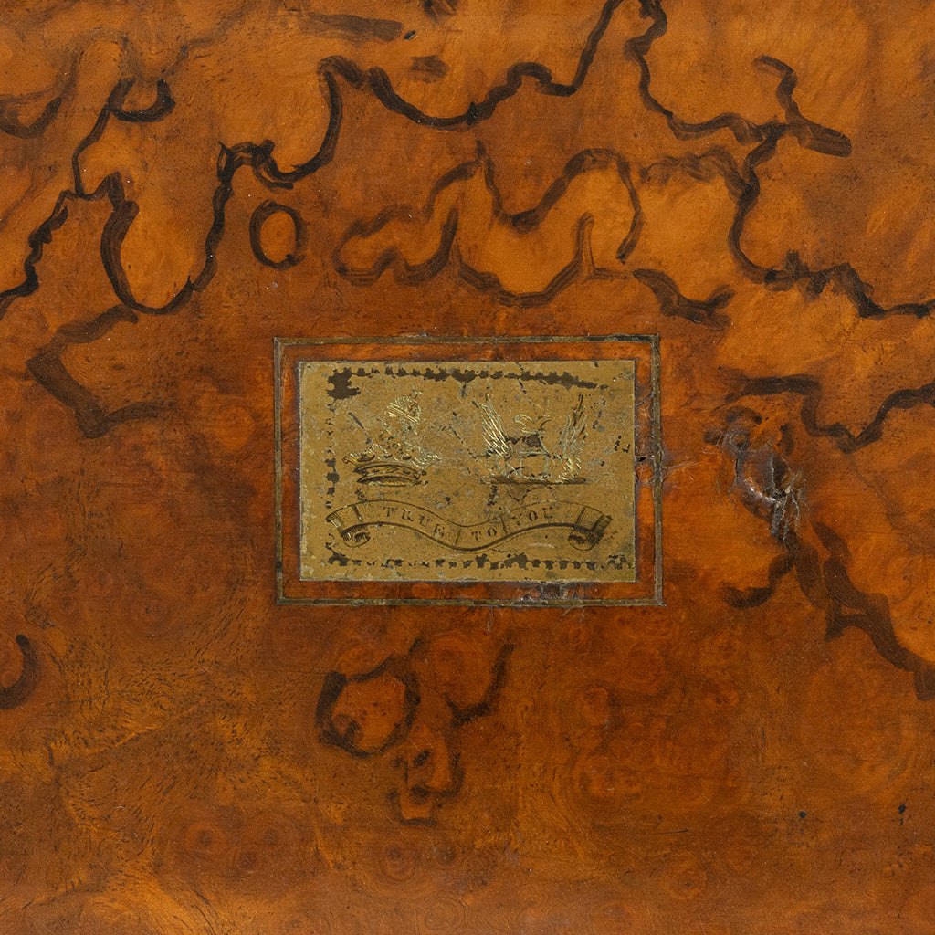 An antique wooden box lid featuring a rich, swirling burl wood pattern in warm brown and orange tones, with a central rectangular metal inset showing a vintage illustration and the text 'true to you'.