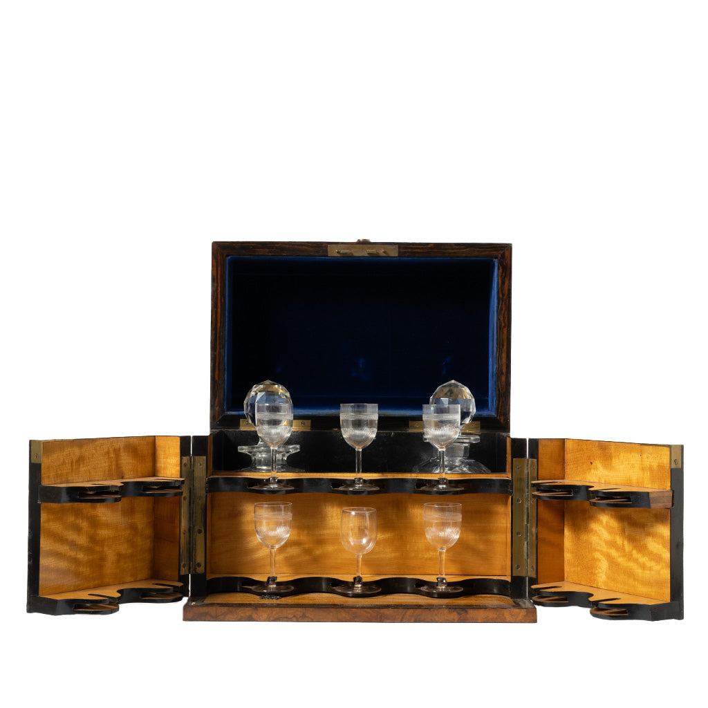 An open burl wood liquor cabinet with a blue velvet-lined interior displays a cut crystal decanter and six matching etched stemmed glasses secured in custom fitted holders. The fold-out side compartments and mirrored back create an elegant presentation while providing organized storage for the glassware.