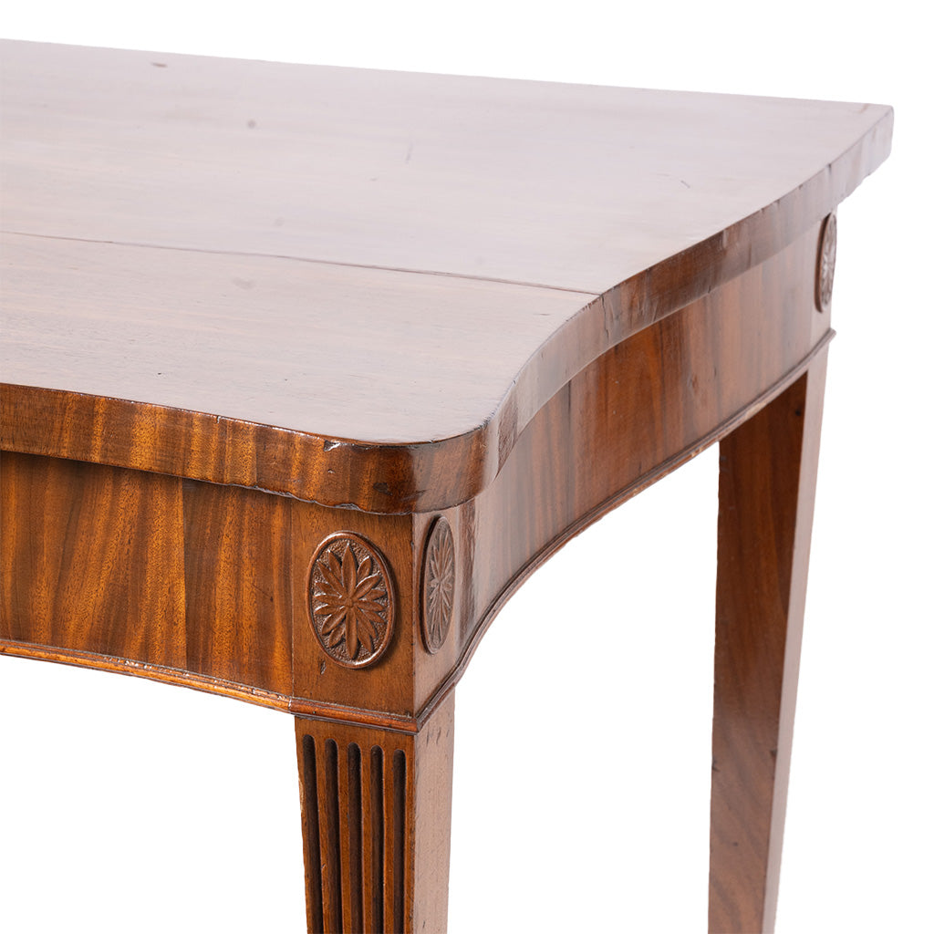 Close-up of a wooden console table corner showing the curved apron, carved circular medallion details, fluted leg, and warm medium-brown wood grain against a white background.