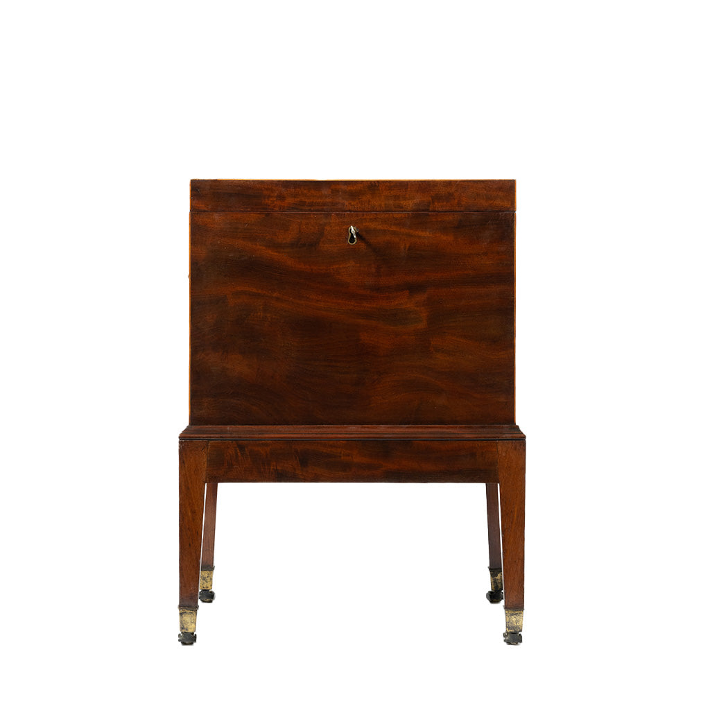Front view of an antique mahogany cellaret with a richly figured wood grain, a central keyhole, and a clean, rectangular silhouette. The piece stands on straight tapered legs with brass-capped casters, reflecting its elegant early 19th-century design.