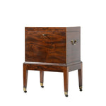 A three-quarter view of an antique mahogany cellaret shows its richly figured wood grain, brass side handles, and central keyhole on the front. The piece stands on straight tapered legs with brass-capped casters, combining elegant craftsmanship with practical storage.