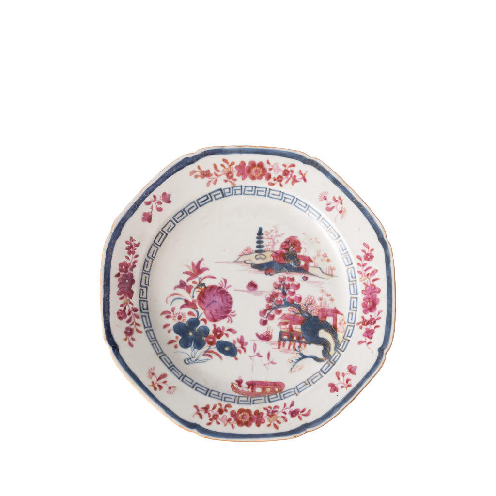 A decorative ceramic plate with a softly scalloped rim features a blue geometric border, pink floral accents, and a thin gold-trimmed edge. The center is adorned with stylized flowers, birds, and a pagoda-inspired landscape in shades of pink and blue, creating a classic chinoiserie-inspired design.