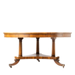 A round wooden table with turned pedestal legs and curved feet on casters, shown against a plain white background.