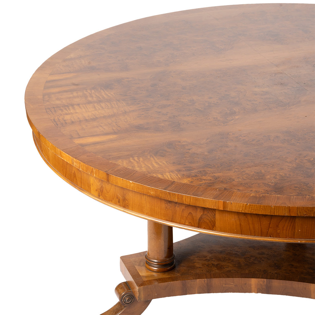 Close-up of a round wooden table with a rich burl wood grain finish, featuring a pedestal base and a lower shelf, shown against a plain white background.