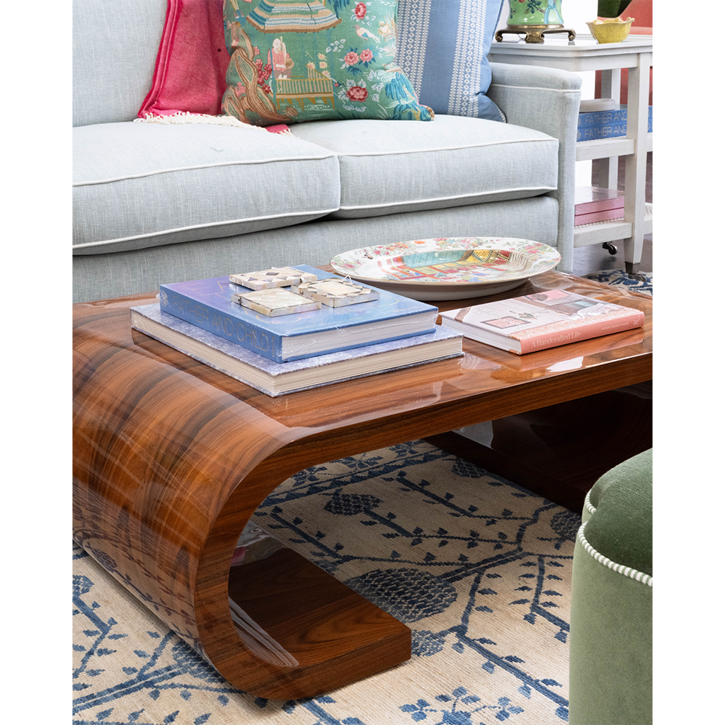 A curved wooden coffee table with a glossy dark brown finish, styled in a living room setting with books, a decorative plate, and a light blue sofa in the background.