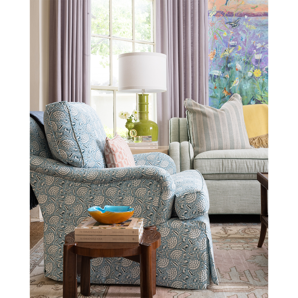A styled living room scene featuring a blue and white patterned slipcovered armchair in the foreground, a matching armchair in the background, a green table lamp, a wooden side table with books and a bowl, and a colorful floral painting on the wall.