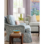 A styled living room scene featuring a blue and white patterned slipcovered armchair in the foreground, a matching armchair in the background, a green table lamp, a wooden side table with books and a bowl, and a colorful floral painting on the wall.