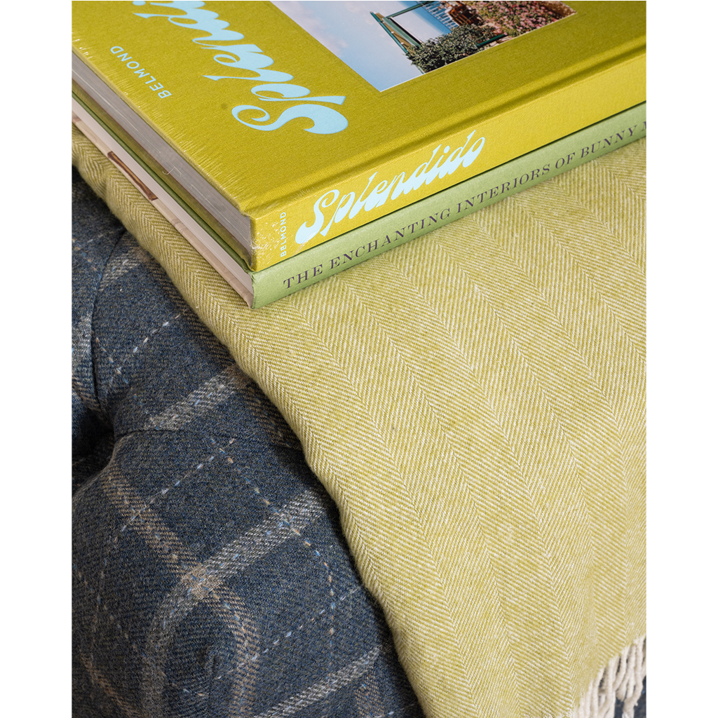 A close-up of a textured herringbone throw blanket in a muted olive-yellow color, styled on a dark gray plaid sofa with two books stacked on top.