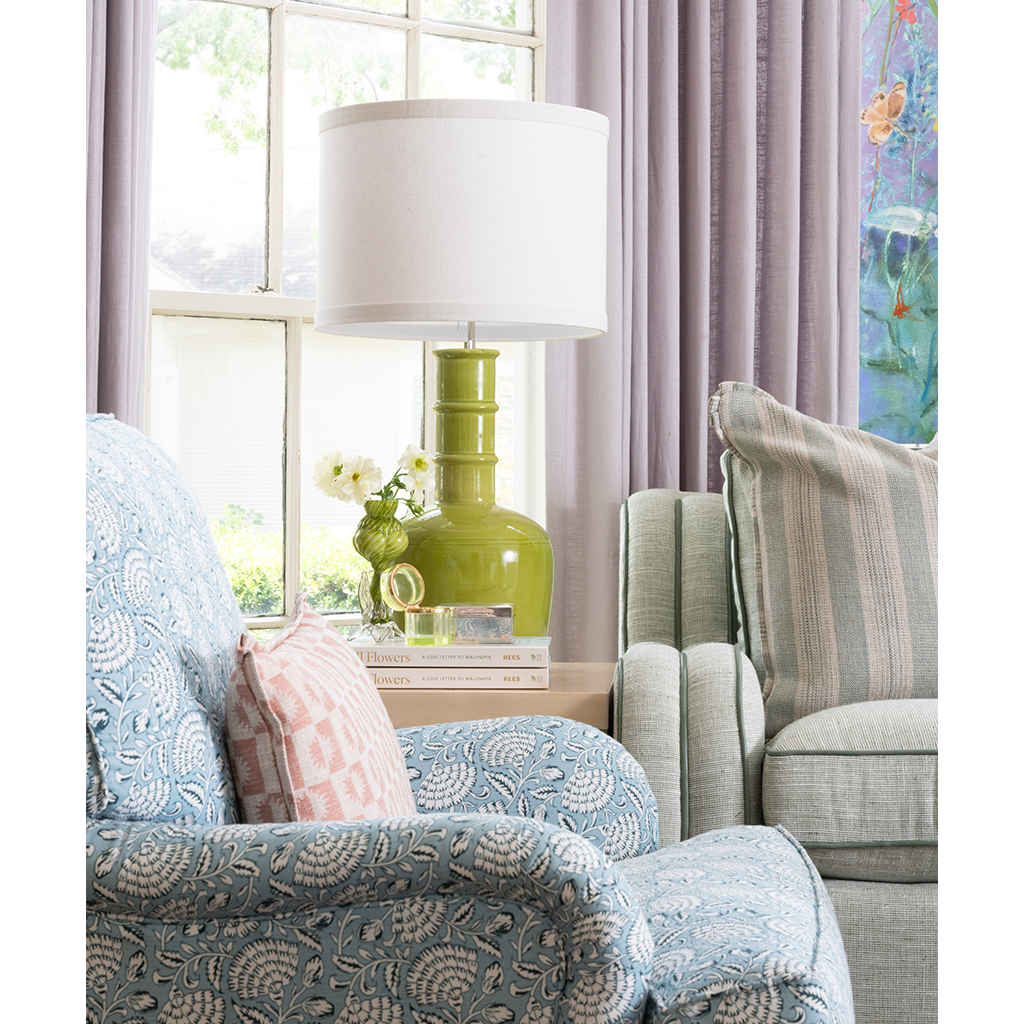 A styled living room corner featuring a light blue floral armchair, a sage green armchair with a striped pillow, and a green ceramic table lamp with a white drum shade on a window table.