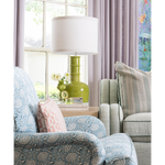 A styled living room corner featuring a light blue floral armchair, a sage green armchair with a striped pillow, and a green ceramic table lamp with a white drum shade on a window table.