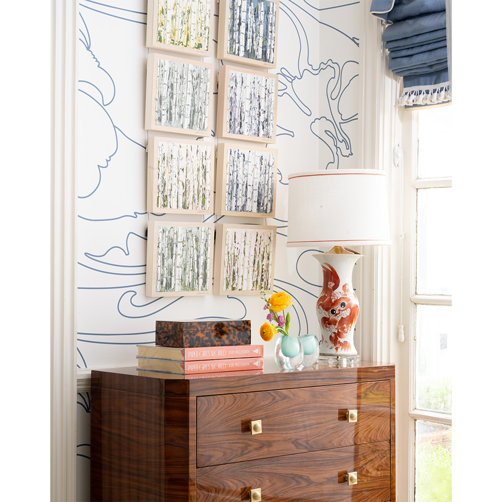 A styled interior corner showing a dark wood dresser against a white wall with blue line art wallpaper, framed birch tree prints, a white table lamp, and decorative objects on top.