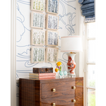 A styled interior corner showing a dark wood dresser against a white wall with blue line art wallpaper, framed birch tree prints, a white table lamp, and decorative objects on top.