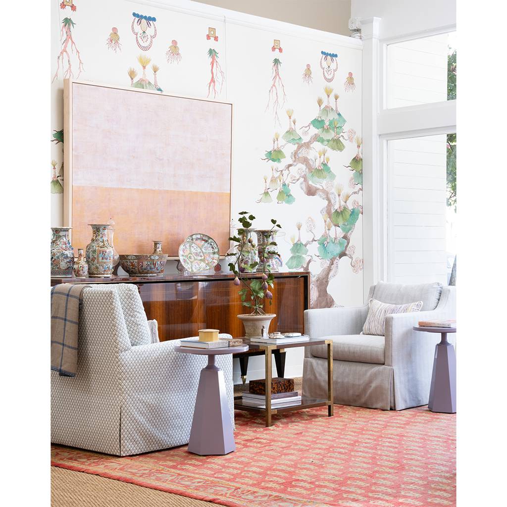A styled living room interior featuring two upholstered armchairs, small side tables, a wooden credenza with decorative vases and plates, a patterned red rug, and a large abstract wall art piece against a white wall with botanical mural wallpaper.