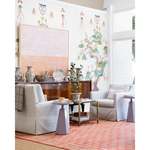 A styled living room interior featuring two upholstered armchairs, small side tables, a wooden credenza with decorative vases and plates, a patterned red rug, and a large abstract wall art piece against a white wall with botanical mural wallpaper.