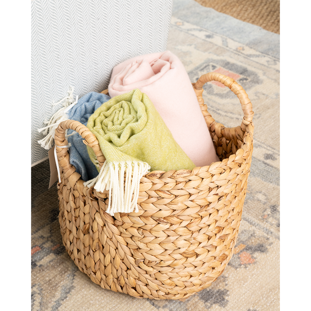 A natural woven seagrass basket with two handles, filled with rolled throw blankets in pink, light green, and blue, set on a patterned rug next to a textured white chair.