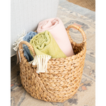 A natural woven seagrass basket with two handles, filled with rolled throw blankets in pink, light green, and blue, set on a patterned rug next to a textured white chair.