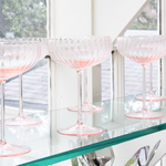 Pink glass goblets on a reflective surface with a blurred window background