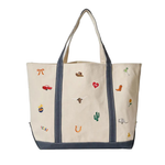 An off-white canvas tote bag with navy blue trim and handles, decorated with various embroidered Western-themed motifs including a longhorn, cowboy boots, cactus, and a red heart.