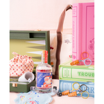 A styled tabletop arrangement of vintage-inspired game accessories and books: an olive green backgammon board set, pink and blue game books, a small pink patterned drawstring bag, a clear bottle with a red-and-blue Christmas label reading 'Merry Christmas,' orange-handled scissors, dice, and a light blue patterned dish.