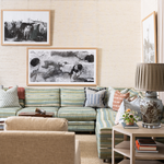 A styled living room featuring a large framed black-and-white art print on a textured beige wall above a green and blue striped sectional sofa. The room includes a matching armchair, a side table with a patterned ceramic lamp and books, and additional framed wall art.