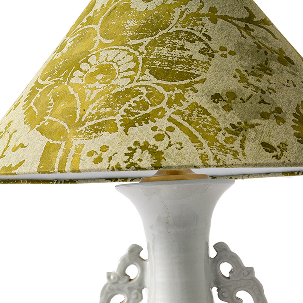 A close-up of a table lamp featuring a textured olive green and cream damask patterned lampshade and a glossy white ceramic base with ornate side details.