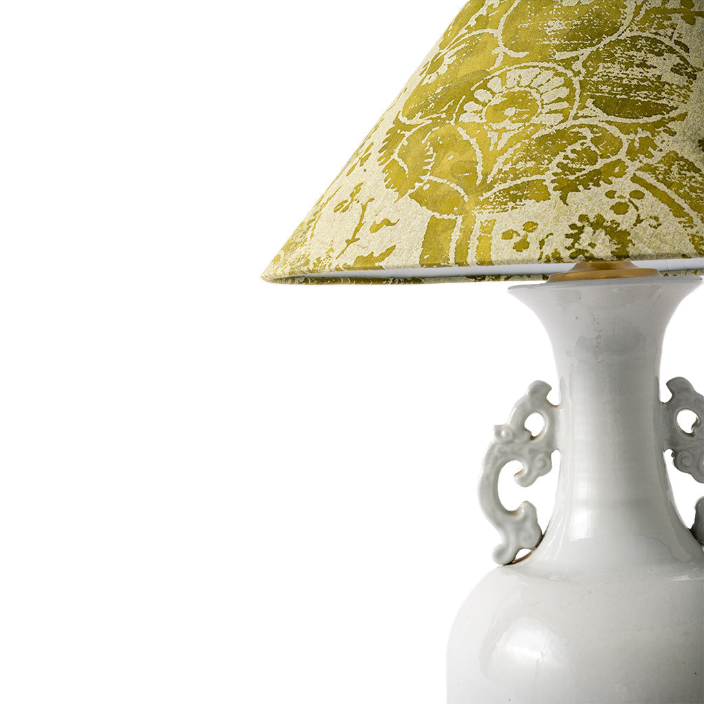 Close-up of a table lamp with a white ceramic body and ornate side handles, featuring a tapered lampshade in a distressed olive green and cream floral pattern, set against a plain white background.